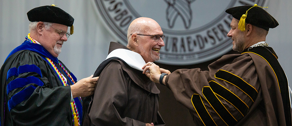 Dr. Jeff Gingerich, university president (right), and Dr. Joseph Zimmer, provost and vice president for academic affairs, honor Fr. Dan Riley, ofm, with an honorary doctorate.