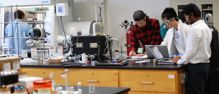 Students work with a professor, surrounded by chemistry equipment.