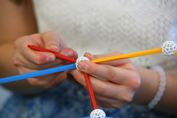 A white Zometool node is the same kind of polyhedron as the modules. Close up of a student's hands as she attaches struts to a node.