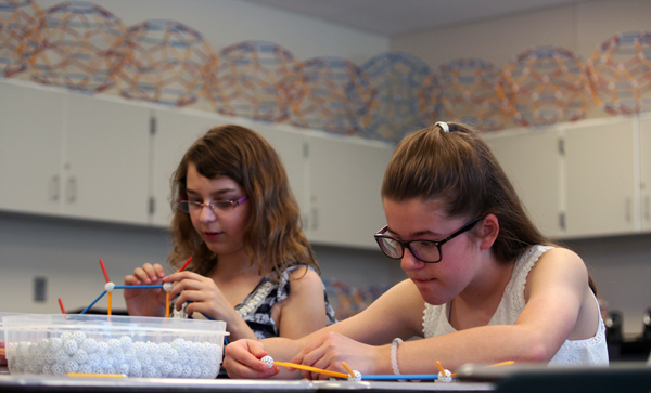 Later classes were surrounded by the fruits of the labors of the earlier classes. Two students build modules. In the background, shelves are filled with modules completed by earlier classes.