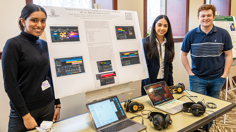 Tanvi, Yasmine, & Joseph stand next to their poster and a table with laptops and headphones.