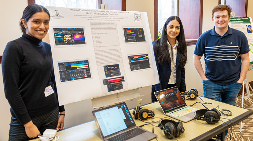 Tanvi, Yasmine, & Joseph stand next to their poster and a table with laptops and headphones.
