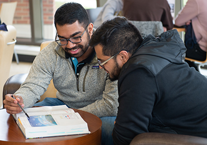 A student tutoring another while looking at an open book.